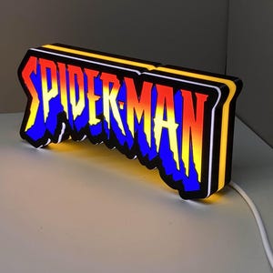 Może przedstawiać: Prostokątny znak LED z napisem SPIDER-MAN stylizowaną, poszarpaną czcionką. Litery mają gradient od żółtego u góry, przez czerwony w środku, do niebieskiego na dole, z czarnym obrysem i świecącą na żółto krawędzią boczną.