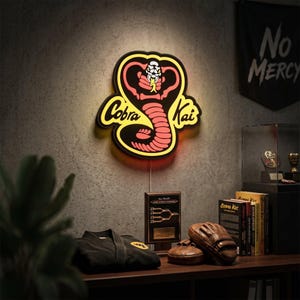 May include: A wall-mounted LED sign featuring the Cobra Kai logo with a stylised cobra snake in red and black, illuminated by a yellow glow. The sign displays the text Cobra Kai in a bold, black script font against a bright yellow background.