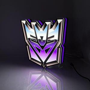Könnte beinhalten: Ein Decepticon-Logo-Wandschild mit einem metallischen Silber- und Violett-Farbverlauf. Das Emblem hat ein scharfes, kantiges Design mit einem leuchtenden violetten Licht, das von den Kanten ausgeht und einen lebendigen Kontrast erzeugt.