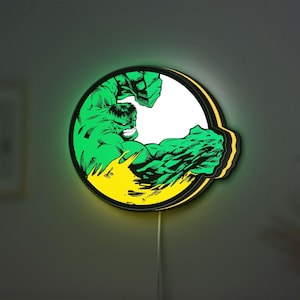 May include: A circular wall-mounted LED light featuring a graphic illustration of the Hulk in a dynamic pose. The character is depicted in vibrant green with black line details, set against a white background with a yellow base, emitting a soft glow.