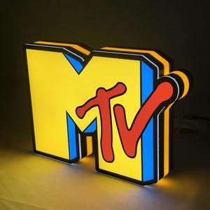 Może przedstawiać: Trójwymiarowy, podświetlany znak z logo MTV, przedstawiający dużą żółtą literę M z niebieskimi akcentami oraz wyrazisty czerwony napis TV. Znak posiada grubą czarną obwódkę i emituje ciepłe światło z krawędzi, tworząc żywy efekt wizualny.