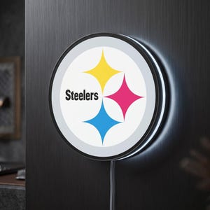 Peut inclure: Une enseigne murale LED circulaire arborant le logo des Pittsburgh Steelers. Le design comprend le mot Steelers en noir à côté de trois étoiles à quatre branches jaune, rouge et bleue sur fond blanc, le tout entouré d'un cadre noir.
