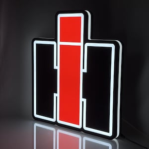 Può includere: Un'insegna rettangolare con il logo International Harvester caratterizzata da una barra rossa verticale centrale affiancata da due pannelli neri, il tutto delineato da una luminosa illuminazione a LED bianca dal design geometrico.