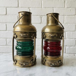 May include: Two brass nautical lanterns with cylindrical bodies and metal handles. One lantern features a green glass center section, while the other lantern features a red glass center section. Both lanterns have a metallic finish and a structured frame.