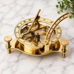 Puede incluir: Un reloj de sol con brújula de latón pulido que presenta una base circular con tres tornillos de nivelación ajustables. El dispositivo incluye un gnomon central, una escala de latitud curva numerada y números romanos.