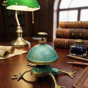 Puede incluir: Un timbre de escritorio de lat&oacute;n con forma de rana y acabado de p&aacute;tina verde sobre la c&uacute;pula y el cuerpo. El timbre presenta un patr&oacute;n texturizado similar a escamas en la c&uacute;pula superior y cuatro patas extendidas. Un peque&ntilde;o pulsador se sit&uacute;a en el centro.