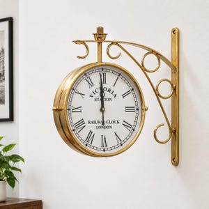 Puede incluir: Un reloj de pared de doble cara con marco y soporte de metal dorado pulido. La esfera blanca presenta números romanos negros y el texto Victoria Station Railway Clock London con manecillas negras que indican la hora.