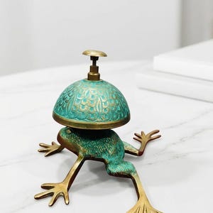 Puede incluir: Un timbre de escritorio de lat&oacute;n con forma de rana y acabado de p&aacute;tina verde. La rana tiene una piel texturizada y patas extendidas, que sostienen una campana en forma de c&uacute;pula con un intrincado patr&oacute;n de escamas grabado y un bot&oacute;n.