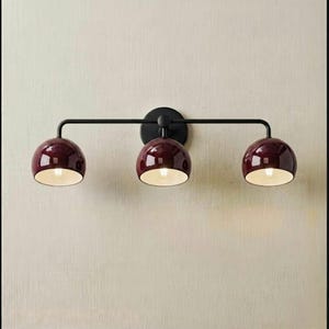 Puede incluir: Una lámpara de pared con una barra horizontal de metal negro y tres pantallas en forma de cúpula con un acabado en color burdeos intenso. Cada pantalla tiene un interior blanco y está orientada para dirigir la luz hacia abajo desde el soporte.