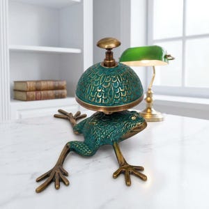 Puede incluir: Un timbre de escritorio decorativo con una base de rana esculpida en acabado verde oscuro y bronce. La c&uacute;pula del timbre presenta un patr&oacute;n texturizado similar a escamas en tonos verde azulado y bronce, rematado con un peque&ntilde;o bot&oacute;n redondo.