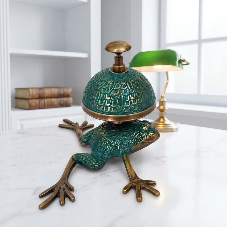 Puede incluir: Un timbre de escritorio decorativo con una base de rana esculpida en acabado verde oscuro y bronce. La c&uacute;pula del timbre presenta un patr&oacute;n texturizado similar a escamas en tonos verde azulado y bronce, rematado con un peque&ntilde;o bot&oacute;n redondo.