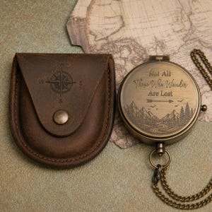 May include: A round brass compass with a chain and a brown leather pouch. The compass lid is engraved with the text Not All Those Who Wander Are Lost, an arrow, and a mountain landscape. The leather pouch features an embossed compass rose design.