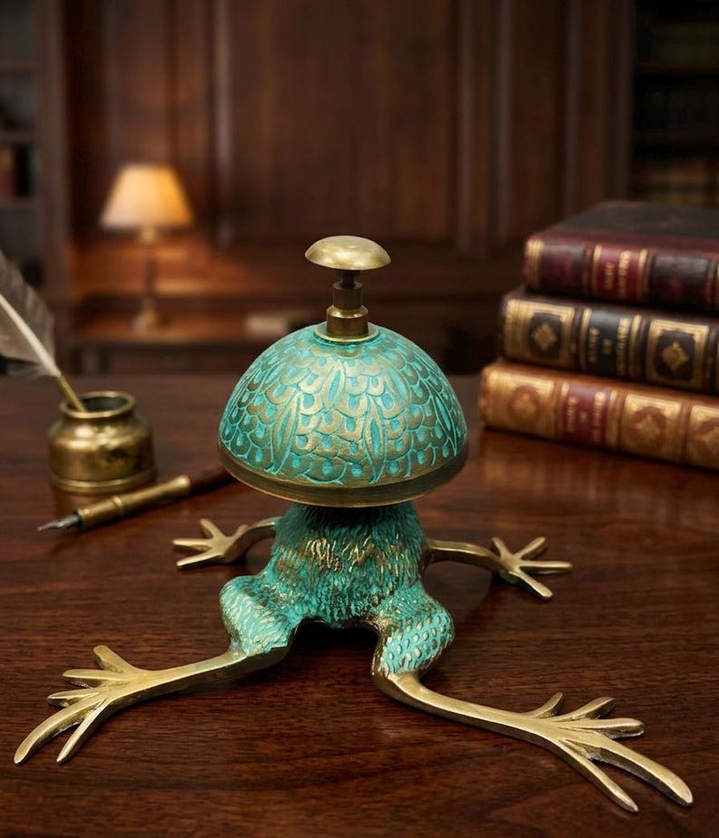 Puede incluir: Un timbre de escritorio con una base en forma de rana con patas extendidas y una parte superior en forma de c&uacute;pula. El objeto de metal tiene un acabado desgastado en color verde azulado y dorado. La c&uacute;pula tiene un patr&oacute;n de plumas.