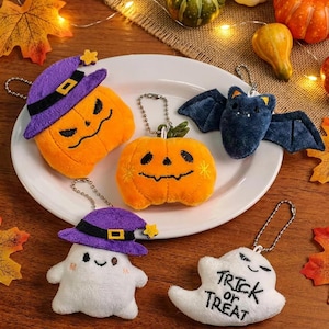 Könnte beinhalten: Fünf Halloween-Plüsch-Schlüsselanhänger auf einem weißen Teller: ein orangefarbener Kürbis mit lila Hexenhut, ein kleiner Kürbis, eine dunkelblaue Fledermaus, ein weißes Gespenst mit lila Hut und ein weißes Gespenst mit der Stickerei Trick or Treat.