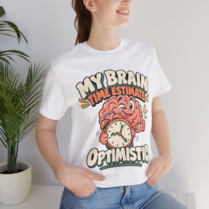 Può includere: Una t-shirt bianca a maniche corte con la grafica di un cervello rosa che tiene in mano una sveglia arancione. Il testo MY BRAIN TIME ESTIMATE OPTIMISTIC è scritto in un font retrò. Il design utilizza colori arancione, crema e verde scuro.