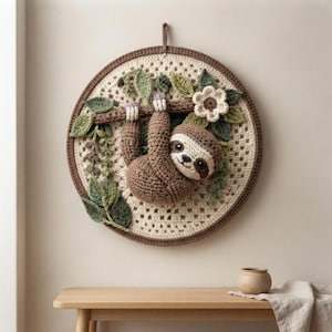 May include: A circular crochet wall hanging featuring a brown sloth hanging from a branch. The background is a cream-colored granny square pattern. The sloth is surrounded by crocheted green leaves and a single white flower with a brown center.