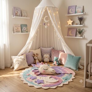 Puede incluir: Un dosel de tela blanca con luces cuelga sobre una alfombra de ganchillo con una cabeza de drag&oacute;n en tonos pastel, estrellas y una luna creciente. Varios peluches, como un ciervo, un b&uacute;ho y un drag&oacute;n, descansan entre cojines cuadrados de colores.