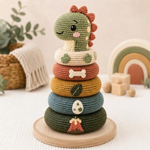 May include: A crochet stacking toy featuring a green dinosaur head topper with a rust-colored spine. The stack consists of four rings in cream, rust, mustard, and dark green, decorated with a white bone, a green leaf, a speckled egg, and a volcano.