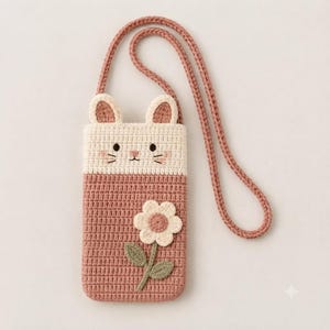 May include: A crochet crossbody bag featuring a cat face design with cream-colored ears and a face on a dusty rose body. The bag includes a long matching strap and a decorative cream flower appliqué with a pink center and green leaves on the front.