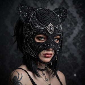 May include: A black crocheted balaclava featuring cat-like ears, intricate silver metallic embroidery, and a central silver-toned filigree ornament with a black gemstone. Small silver key charms hang from the lower edge of the face opening.