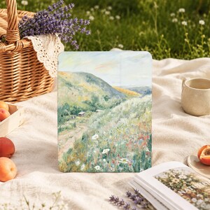 Puede incluir: Una funda rectangular para tableta con una pintura paisajística impresionista de una colina verde, un camino de tierra y un campo de flores silvestres en tonos blancos, amarillos y rosas bajo un cielo pálido y montañas lejanas.