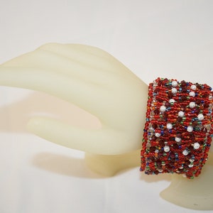 May include: A red, white, and blue beaded bracelet with a geometric pattern. The bracelet is made with small seed beads and larger round beads.