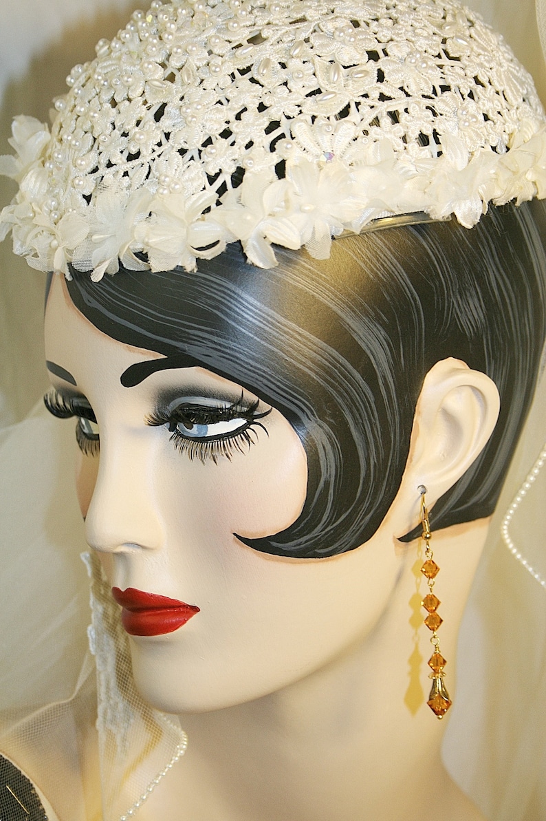 May include: A white crocheted hat with pearl accents and a white flower trim. The hat is worn by a mannequin with black hair and a pair of long, amber-colored earrings.