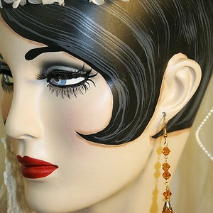 May include: A mannequin head with a white lace headband and long black hair. The mannequin is wearing a pair of gold dangle earrings with amber colored crystals.