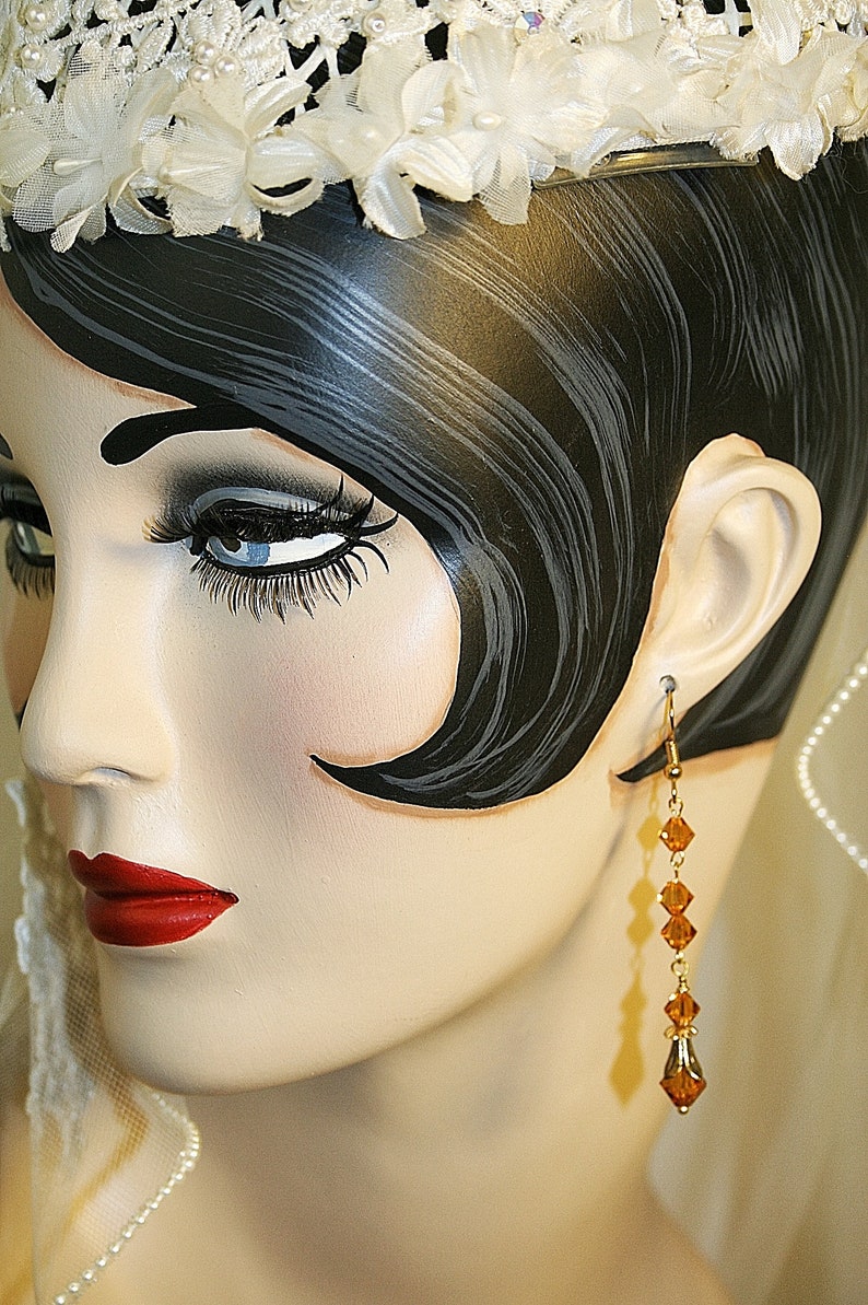 May include: A mannequin head with a white lace headband and long black hair. The mannequin is wearing a pair of gold dangle earrings with amber colored crystals.