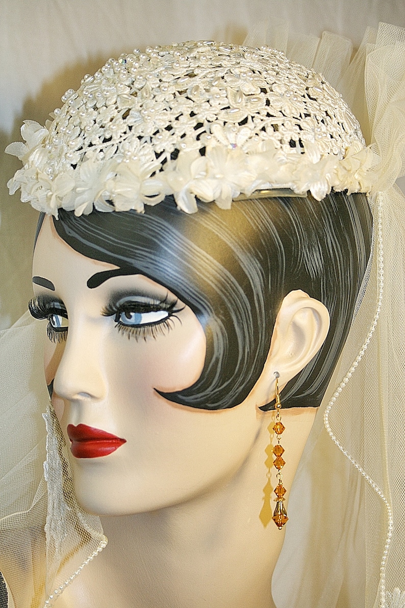 May include: A white crocheted hat with pearl accents and a veil. The hat is decorated with white flowers and is worn by a mannequin with dark hair and long amber earrings.