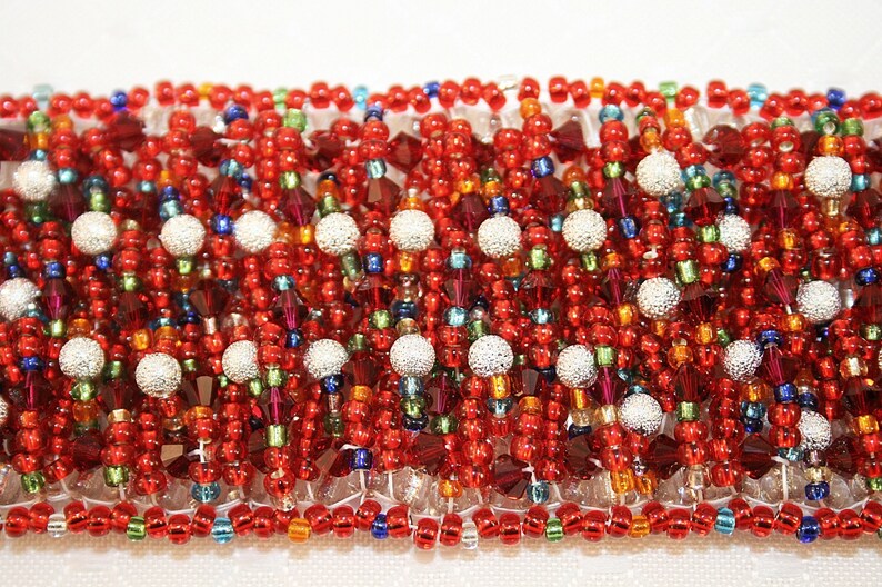 May include: A close-up of a beaded bracelet with a red, orange, green, blue, and silver color scheme. The bracelet is made of small, round beads and has a textured, woven appearance.