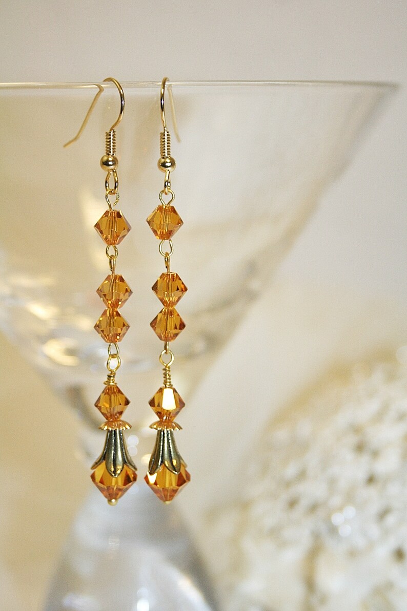 May include: A pair of long gold dangle earrings with amber-colored crystal beads. The earrings have a teardrop-shaped crystal at the bottom.