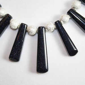 Puede incluir: Un collar con cuentas plateadas y colgantes de piedra preciosa azul oscuro. Las piedras preciosas son facetadas y tienen un acabado brillante.