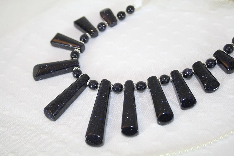 Puede incluir: Un collar hecho con cuentas azules oscuras en forma de estrella y separadores plateados. Las cuentas est&aacute;n dispuestas en forma de abanico, con las cuentas m&aacute;s largas en el centro y las m&aacute;s cortas en los bordes.