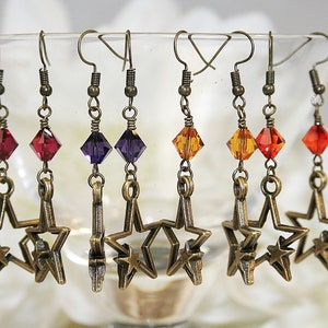 May include: A collection of star-shaped earrings with a vintage bronze finish. Each earring features a colorful, faceted crystal bead in shades of red, purple, and orange. The earrings are displayed against a soft, blurred background.