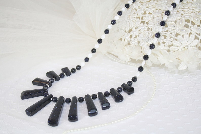 Puede incluir: Un collar largo con cuentas blancas y azules. El collar tiene un gran colgante hecho de varias piedras rectangulares de color azul oscuro.