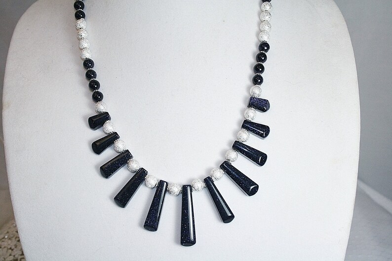 Puede incluir: Un collar de cuentas negras y plateadas con una fila graduada de piedras de color azul oscuro, facetadas, en forma de l&aacute;grima.