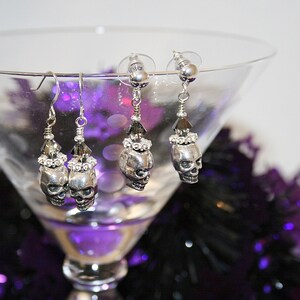 May include: Silver-toned skull earrings with crystal accents. The earrings are displayed in a martini glass with a purple and black background. The earrings have a skull design with crystal embellishments and are perfect for Halloween or gothic-themed outfits.