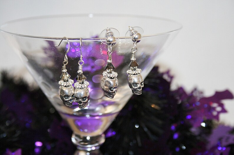 May include: Silver-toned skull earrings with crystal accents. The earrings are displayed in a martini glass with a purple and black background. The earrings have a skull design with crystal embellishments and are perfect for Halloween or gothic-themed outfits.