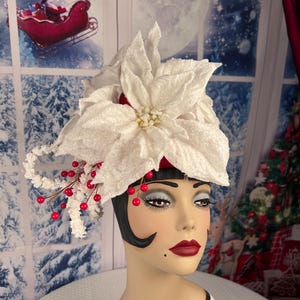 May include: A white poinsettia flower hat with red berries and a decorative head. The hat is adorned with glitter and is set against a winter-themed backdrop. The mannequin head has dark hair, red lipstick, and dramatic eye makeup.