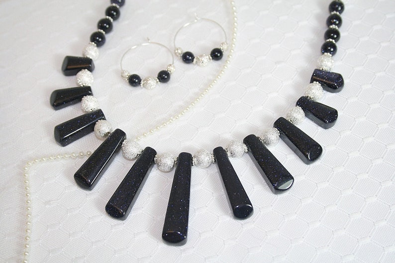Puede incluir: Un collar negro y plateado con un colgante hecho de varias piezas de piedras preciosas azules. El collar se combina con pendientes a juego.