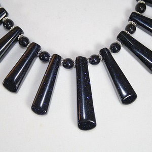 Puede incluir: Un collar hecho de cuentas de piedra preciosa azul oscuro con un acabado brillante. El collar presenta una fila de cuentas m&aacute;s peque&ntilde;as y una fila de cuentas m&aacute;s grandes en forma de l&aacute;grima.