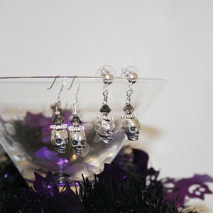 May include: Silver skull earrings with black and clear crystal accents. The earrings are displayed in a martini glass with purple and black Halloween decorations. The skull earrings are a spooky accessory.
