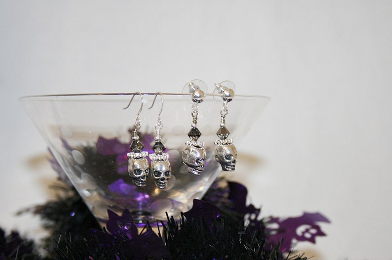 May include: Silver skull earrings with black and clear crystal accents. The earrings are displayed in a martini glass with purple and black Halloween decorations. The skull earrings are a spooky accessory.
