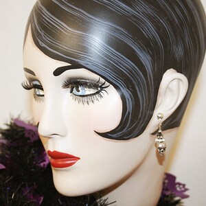 May include: A mannequin head with painted features, including dark hair, long eyelashes, and red lipstick. The mannequin wears a pair of silver earrings with skull charms. A black and purple decorative boa is around the neck.