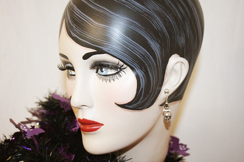May include: A mannequin head with painted features, including dark hair, long eyelashes, and red lipstick. The mannequin wears a pair of silver earrings with skull charms. A black and purple decorative boa is around the neck.