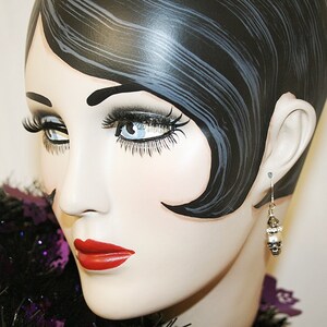 May include: A mannequin head with dark hair and red lipstick is shown wearing a dangling earring. The earring has a silver hook and a cluster of white and silver beads. A black and purple feather boa is around the neck.