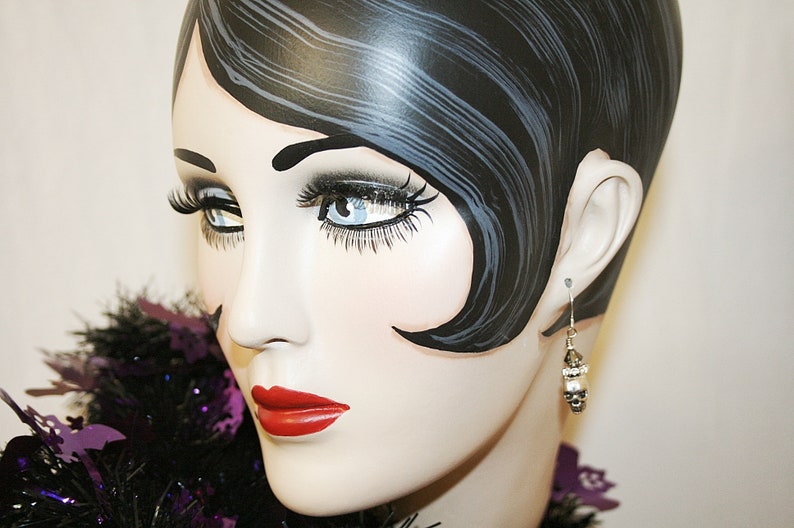 May include: A mannequin head with dark hair and red lipstick is shown wearing a dangling earring. The earring has a silver hook and a cluster of white and silver beads. A black and purple feather boa is around the neck.