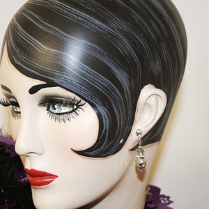 May include: A mannequin head with black hair styled in a vintage wave. The mannequin wears a pair of silver earrings with a pearl-like drop. The mannequin has red lipstick and long eyelashes. A purple feather boa is around the neck.