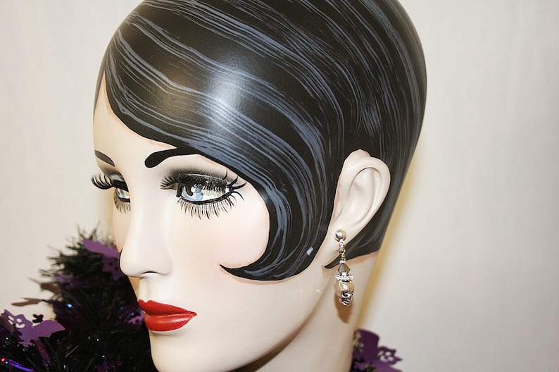 May include: A mannequin head with black hair styled in a vintage wave. The mannequin wears a pair of silver earrings with a pearl-like drop. The mannequin has red lipstick and long eyelashes. A purple feather boa is around the neck.